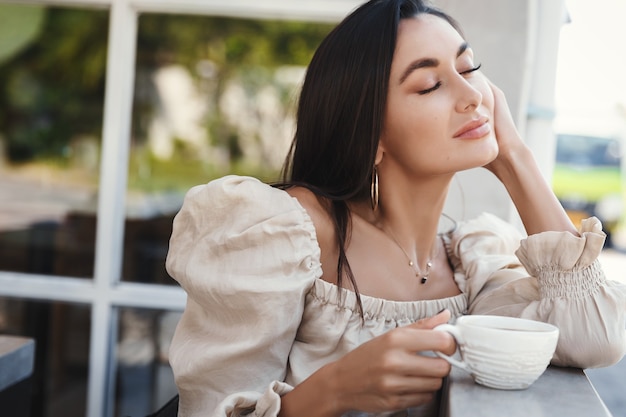 Smiling woman sitting outdoors at hotel restaurant and drinking coffee.