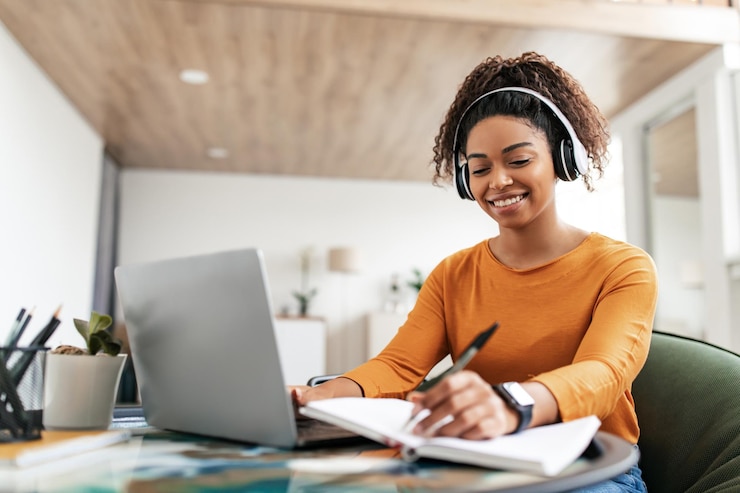 Photo smiling woman sitting at table using computer writing in notebook