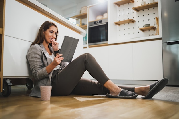 Smiling woman spending leisure time and using digital tablet while sitting on the floor in kitchen at her home.