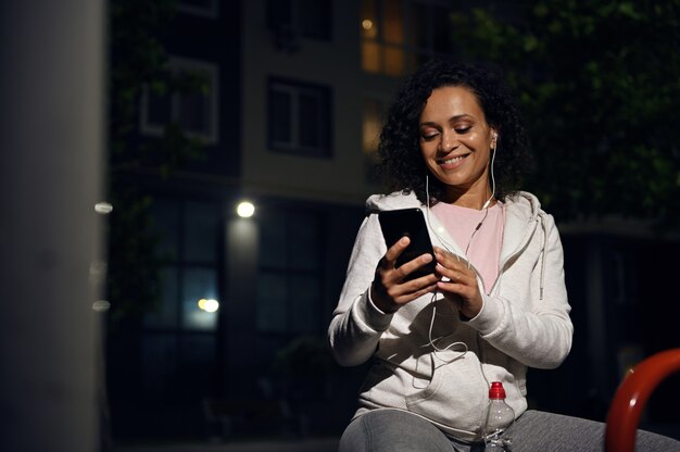 イヤホンを着用し、携帯電話を使用して、照明付きの建物の背景で夜のトレーニングの後に休んで、木製のベンチに座っているスポーツウェアの笑顔の女性