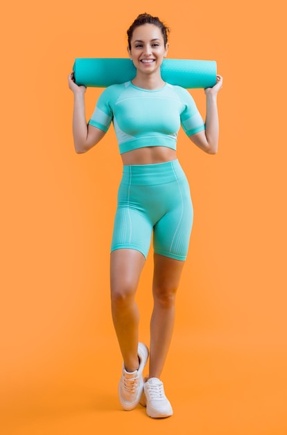 Улыбающаяся женщина в спортивной одежде с фитнес-матом в студии фитнес женщина в спорной одежде держит мат изолированно на желтом фоне фитнес и спорт
