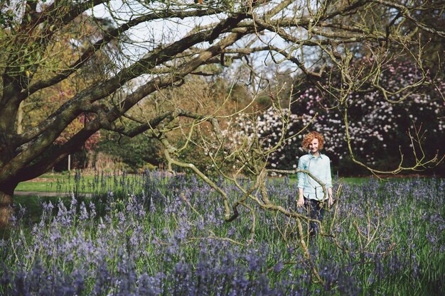 写真 森の木のそばで紫色の花がく植物の真ん中に立っている笑顔の女性