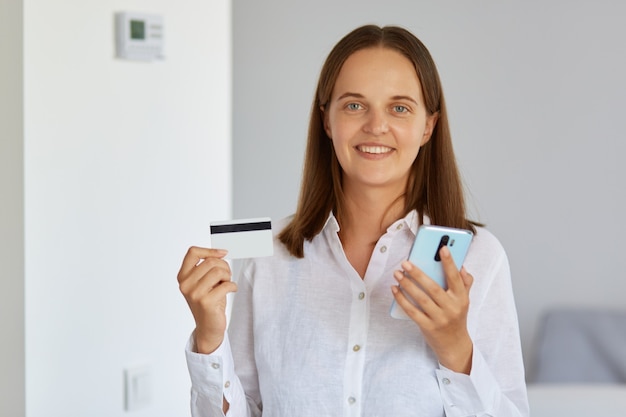 Smiling woman standing indoor with credit card and cell phone in hands, looking at camera, making online payments for shopping in fashion store, wearing white shirt.