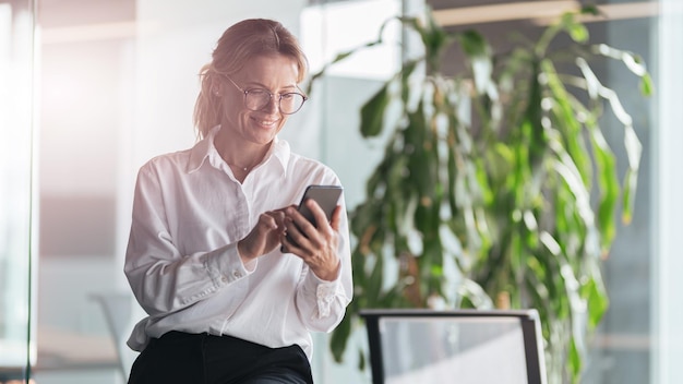 Smiling woman standing in office businesswoman with mobile phone in hand looking at smartphone