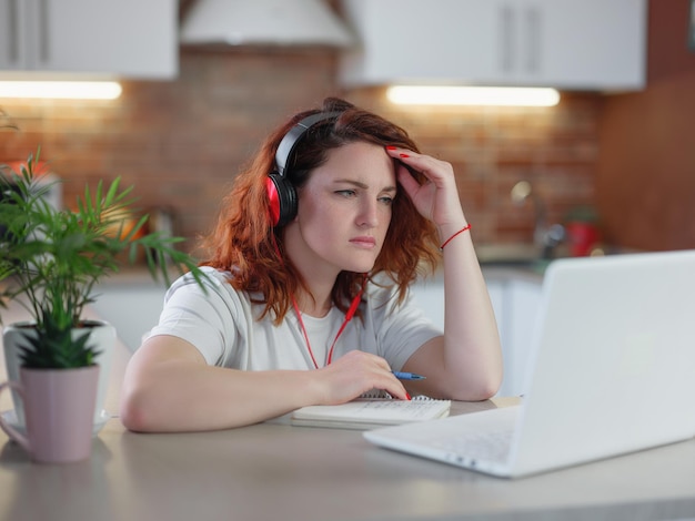 笑顔の女子学生は、彼女の台所でスカイプの先生と一緒に赤いヘッドフォンをオンラインで勉強します