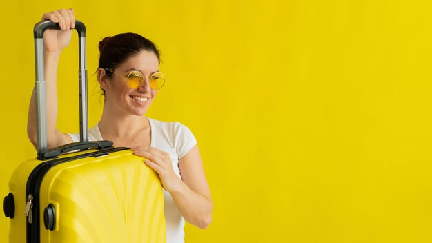 Smiling woman in sunglasses holding suitcase with retractable handle on yellow background