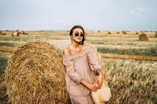 Улыбается женщина в темных очках с открытыми плечами на пшеничном поле и тюки сена