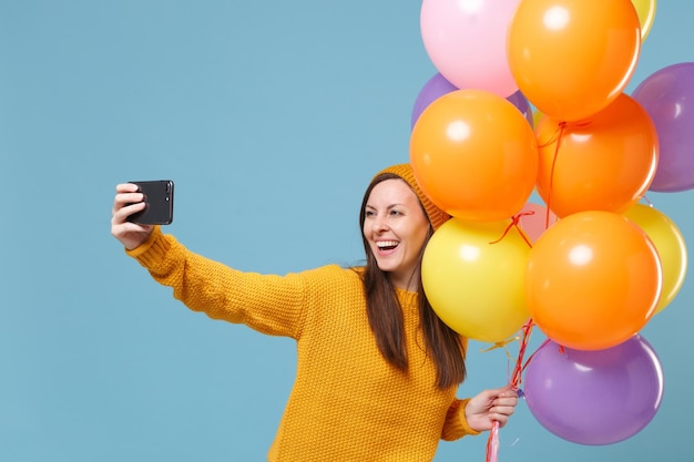 Smiling woman in sweater hat posing isolated on blue background. Birthday holiday party, people emotions concept. Mock up copy space. Celebrating hold air balloons doing selfie shot on mobile phone.