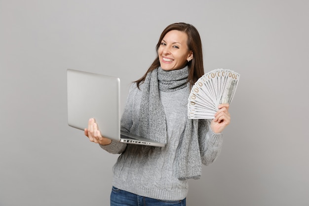 Smiling woman in sweater working on laptop pc computer hold lots bunch of dollars banknotes cash money isolated on grey background. Healthy lifestyle, online treatment consulting, cold season concept.