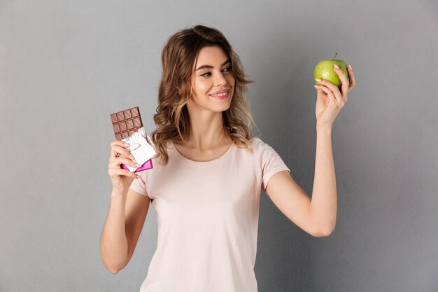 Smiling woman in t-shirt choosing between chocolate and fresh apple over grey