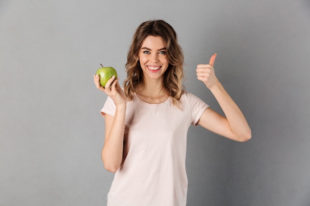 Smiling woman in t-shirt holing fresh apple and showing thumb up while over grey