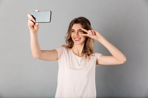 Smiling woman in t-shirt making selfie on smartphone while showing peace gesture over grey