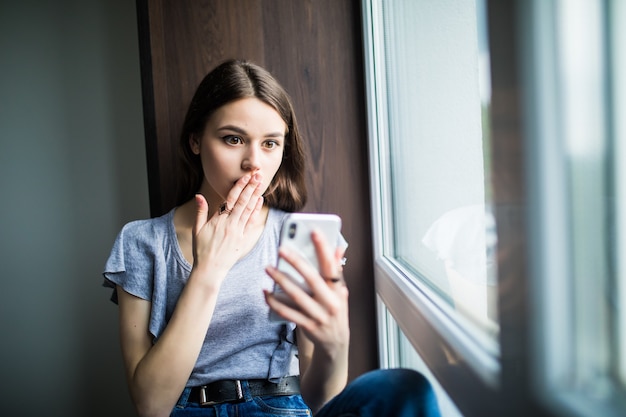 Smiling woman in t-shirt sitting on the windowsill and writing message on smartphone at home