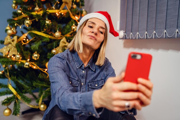 Foto donna sorridente che si fa un selfie mentre è seduta vicino all'albero di natale a casa