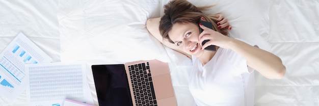 Smiling woman talking on phone and working on laptop while lying on white bed remote work and