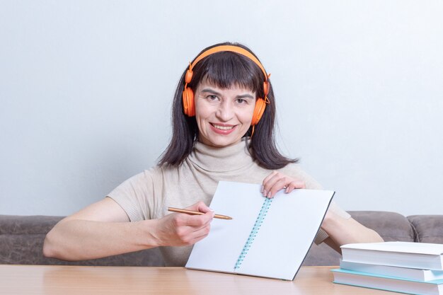 Smiling woman teacher in headphones explains the lesson by showing on a blank exercise book