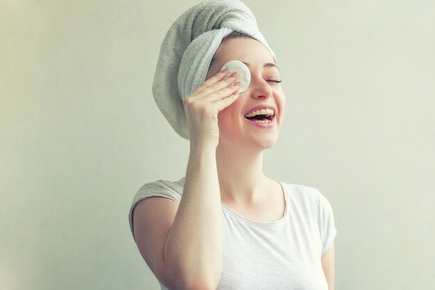 Smiling woman in towel on head with soft healthy skin removing make up with cotton pad isolated on white background