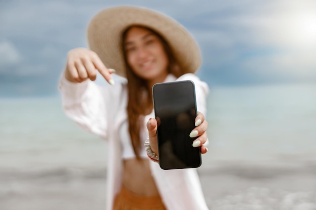 Smiling woman traveler in straw hat showing on phone standing on background of ocean