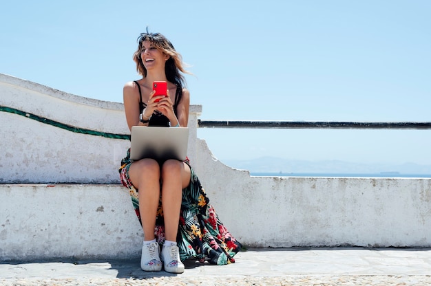 거리에서 노트북으로 작업하는 동안 휴대전화로 타이핑하는 웃는 여자
