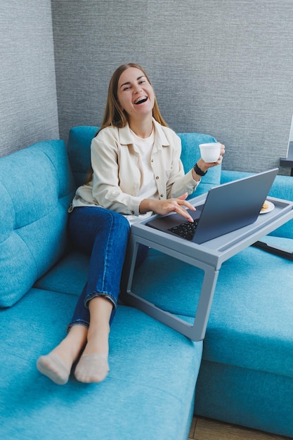 Smiling woman using laptop to check email news online while\
sitting on sofa working on computer writing blog or watching\
webinar studying at home