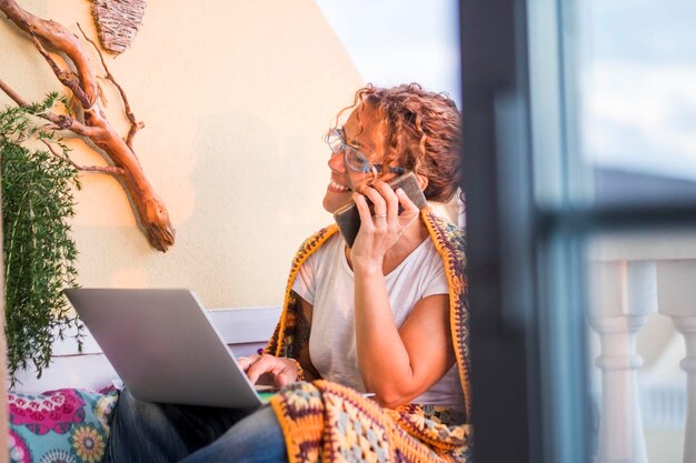 Фото Улыбающаяся женщина использует ноутбук, разговаривая по мобильному телефону дома