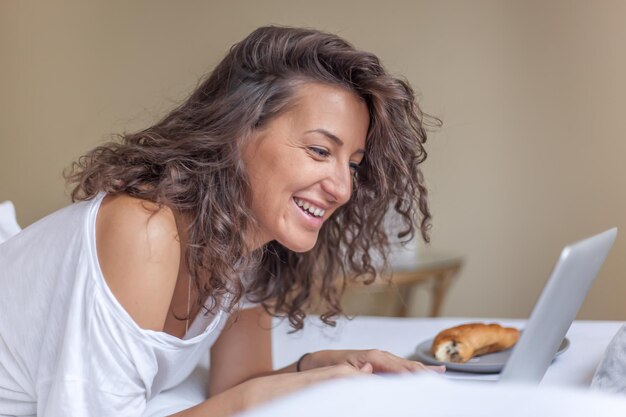 家でベッドで朝食をとってラップトップを使っている笑顔の女性