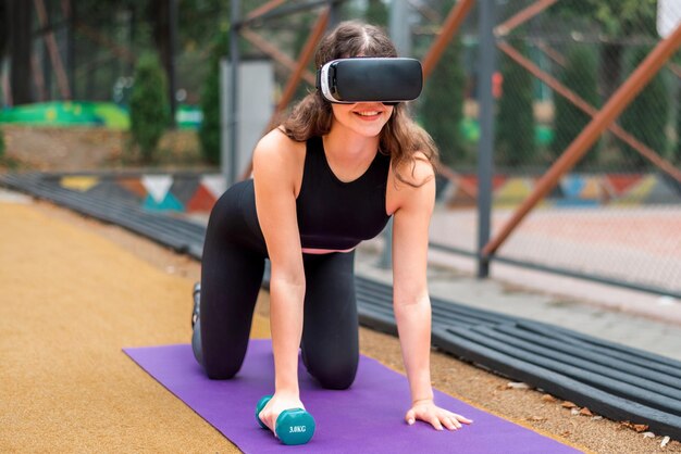 Smiling woman in VR headset in a tracksuit doing exercises with dumbbells on a yoga mat on sports