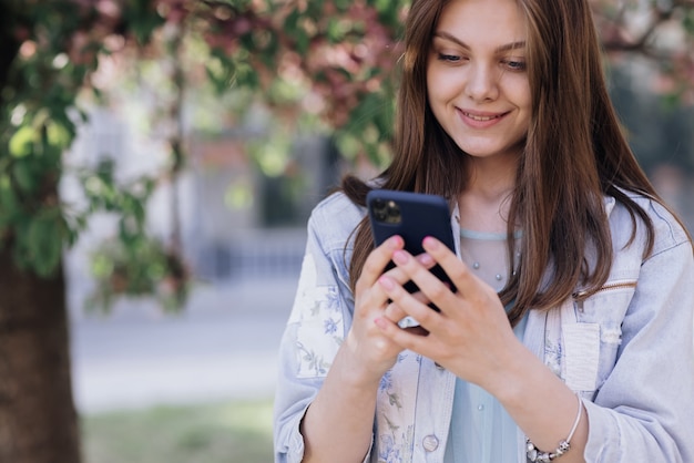 笑顔の女性は通りを歩き、彼女の携帯電話を見て白いドレスを着て彼女の電話を使用します