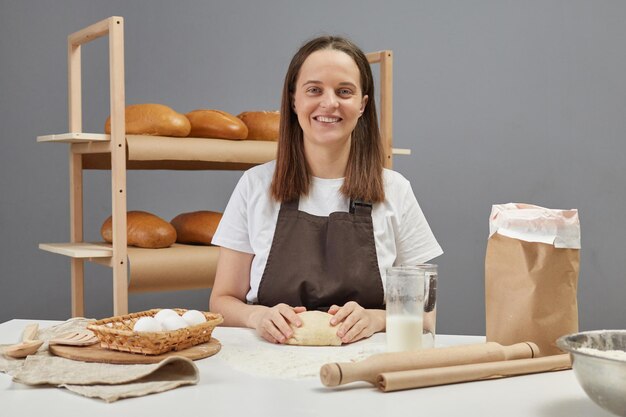 Foto donna sorridente che indossa un grembiule marrone che impasta l'impasto di lievito con la mano per cucinare il pane mentre è seduta