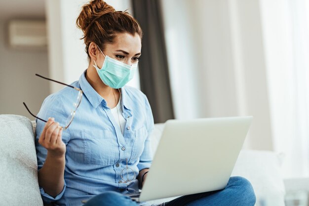 얼굴 마스크를 쓰고 노트북으로 인터넷 서핑을 하고 바이러스가 유행하는 동안 집에서 휴식을 취하는 웃는 여성