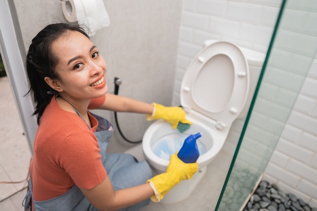 장갑을 끼고 웃는 여자는 화장실에서 화장실을 청소하는 동안 세제 한 병을 보유하고 있습니다.