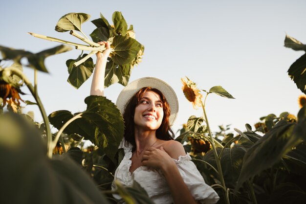 Smiling woman wearing a hat standing in a field of sunflowers in sunset