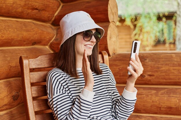 Smiling woman wearing striped shirt panama sunglasses sitting in wooden house terrace having video call waving hand saying hello or bye online communicating internet conference