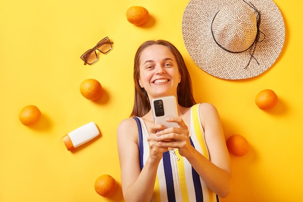 幸せな表情でインターネットを閲覧しながら携帯電話を使用して、黄色の背景にオレンジ色の日焼け止めを塗って横たわっている縞模様の水着を着た笑顔の女性