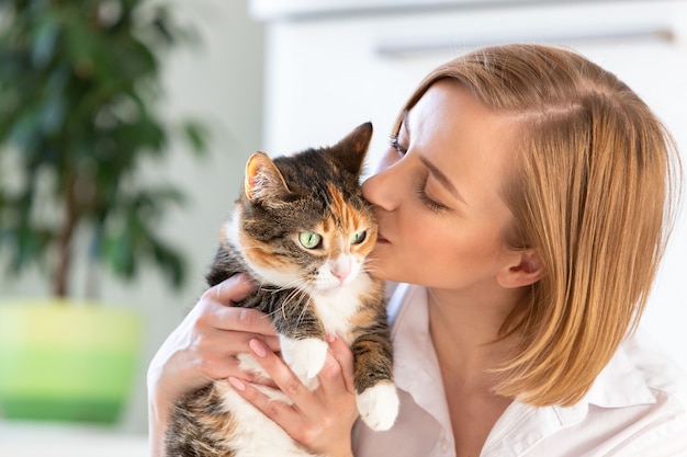 Donna sorridente in camicia bianca che bacia e che abbraccia con tenerezza e amore gatto, tenendola tra le braccia