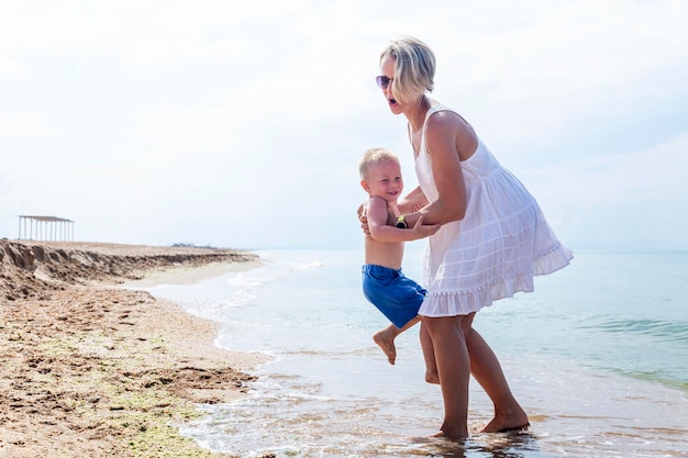A smiling woman in a white sundress with a little boy in blue shorts play on the seashore on a sunny day Love and tenderness Travel and recreation
