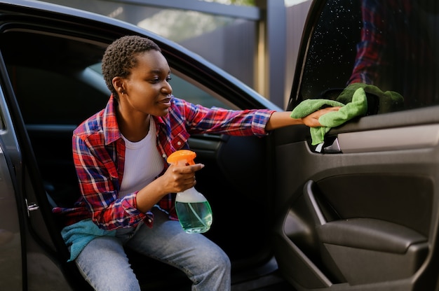 La donna sorridente pulisce un'auto con uno straccio, stazione di lavaggio automatico a mano. industria o attività dell'autolavaggio. la persona di sesso femminile pulisce il suo veicolo dallo sporco all'aperto