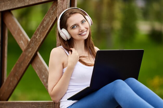 Smiling woman in wireless headphones uses laptop outdoors