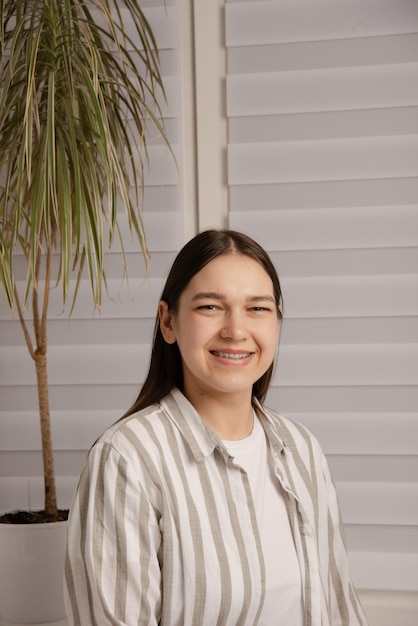 smiling woman with braces looking at camera in room with light closed blinds on window