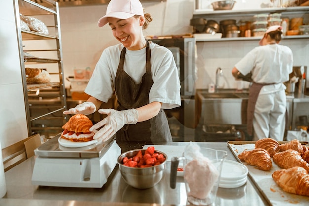 Smiling woman with croissant with strawberry and cream on digital scales in bakery