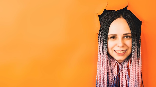 Улыбающаяся женщина с дредами смотрит в камеру Веселая молодая женщина с разноцветными волосами смотрит в камеру в отверстии на оранжевом фоне в студии