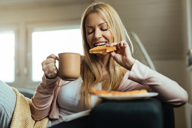 目を閉じてワッフルを食べて、家でコーヒーを飲む笑顔の女性