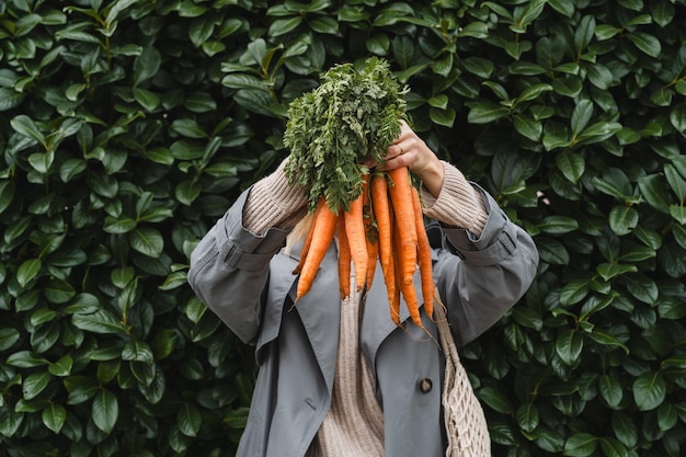 Smiling woman with fresh organic carrots and mesh bag against green leaf wall Concept of people doing sustainable shopping of eco local products without plastic packaging Minimalist lifestyle