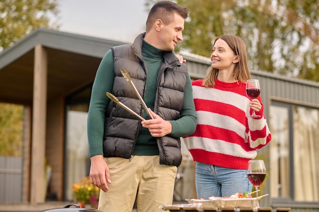 Smiling woman with a glass of wine standing beside the man with the grill tongs