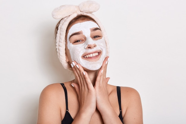 Smiling woman with hairband on her head applied mask to skin of her face, looking smiling directly at camera, keeping palm on her cheeks, posing isolated over white background.