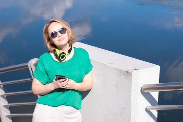 A smiling woman with headphones holding a phone in her hand listens to music standing on the riverbank