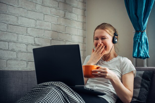 Smiling woman with headphones and laptop on her lap. Young girl on the sofa, holding a cup of tea.