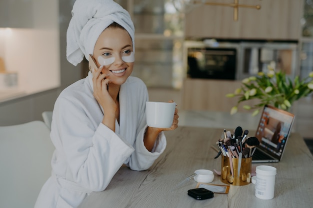 Photo smiling woman with hydrogel patches under eyes enjoys an hot drink tea while speaking on the phone