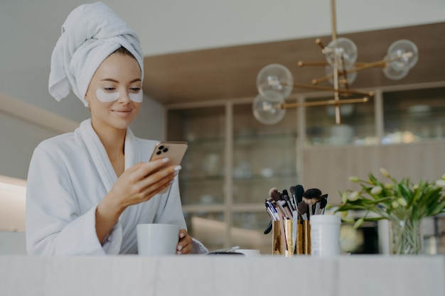 smiling woman with hydrogel patches under eyes enjoys an hot drink tea while using the mobile phone