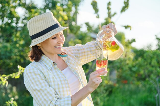 Smiling woman with jug of natural homemade drink in summer garden, herbal tea mint strawberries